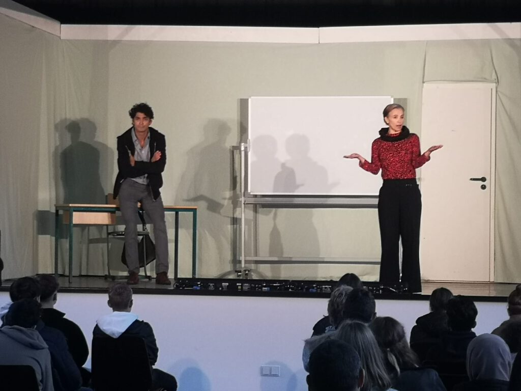 Ein Mann und eine Frau auf der Bühne, es sieht aus wie ein Klassenraum mit Whiteboard (Szenenbild)