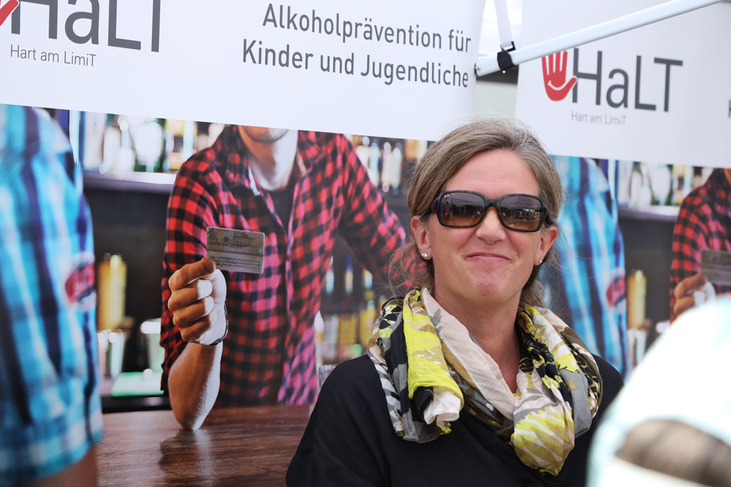 Eine Frau mit Sonnenbrille steht vor einem Aufsteller über dem Steht: Alkoholprävention für Kinder und Jugendliche, daneben ist eine Hand gezeichnet mit der Schrift "HaLT"