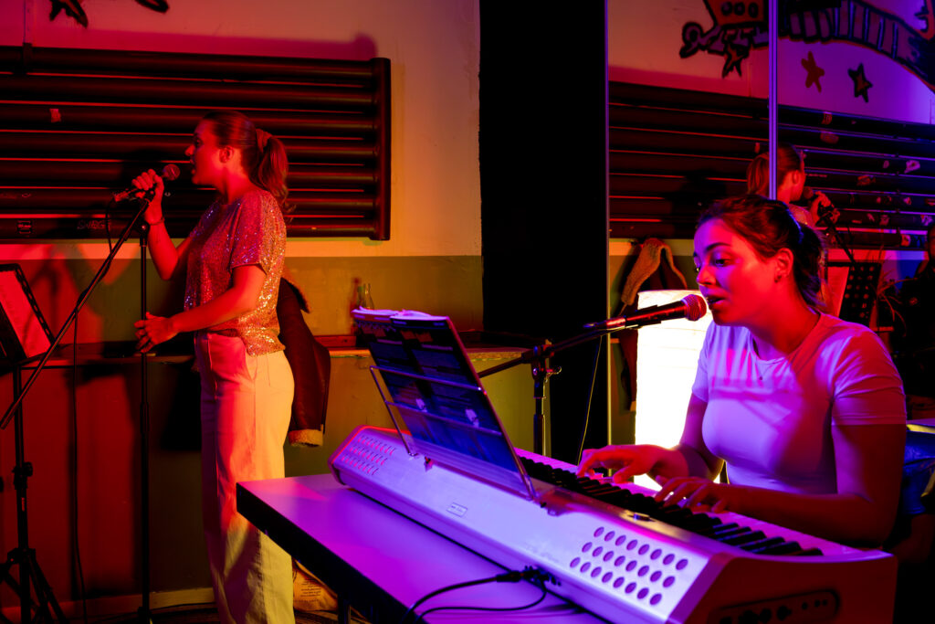 eine junge Frau singt in ein Mikrofon und eine junge Frau spielt Klavier während sie singt.