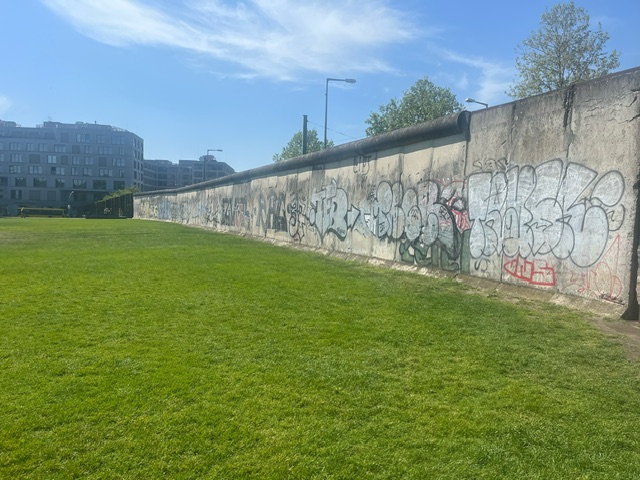 Blick auf den Rest der Mauer mit Grafiti darauf - davor eine grüne Wiese