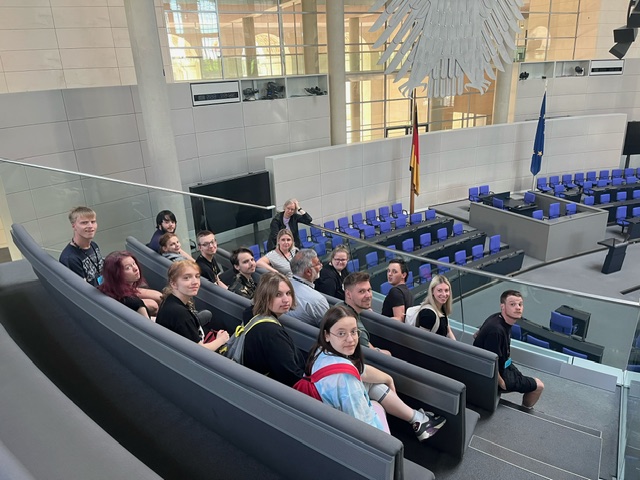 Jugendliche sitzen im Bundestag auf der Tribüne ganz links. Darunter leere Sitze im Plenarsaal, da heute keine Sitzung im Bundestag ist. Die Jugendlichen drehen sich alle um und schauen in die Kamera.